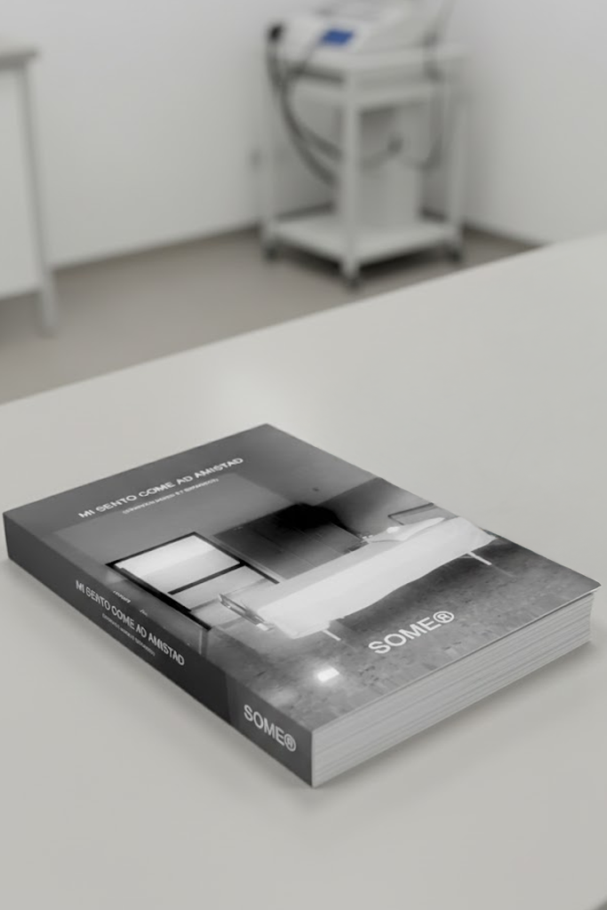 Volume 'MI SENTO COME AD AMISTAD' posizionato in ambiente sterile. Sfondo con strumentazione medica sfocata. SOME® Clinical Archive series.